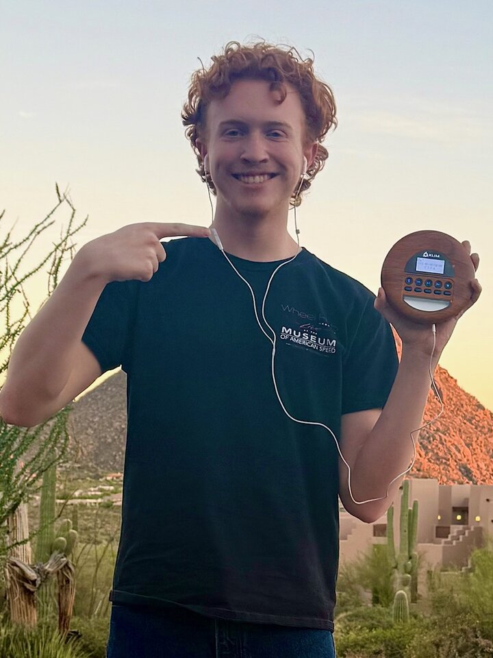 Person holding a music playing device with headphones, standing outdoors near desert plants and a mountain.