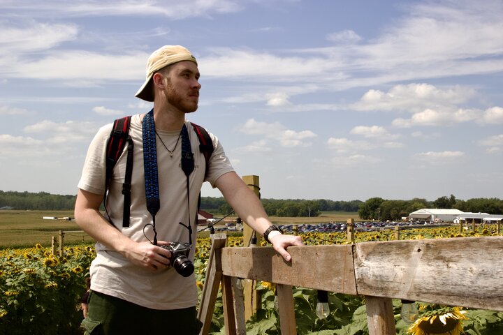Person holding a camera, standing near a wooden fence in a sunflower field under a sunny sky.